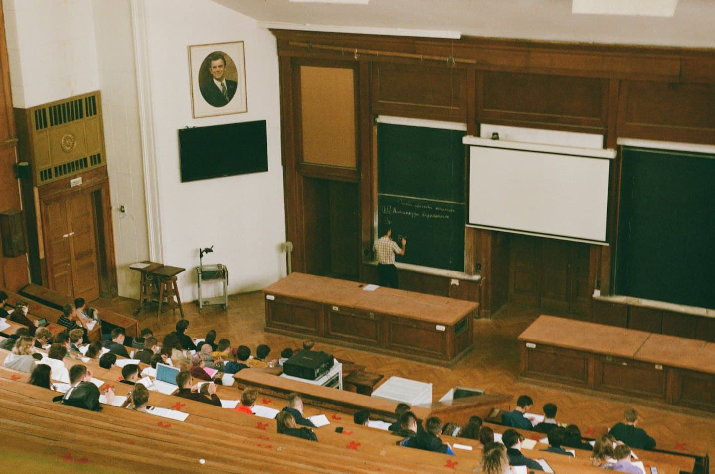 Männlicher Dozent schreibt in einem historischen Hörsaal mit Studenten auf eine Tafel.